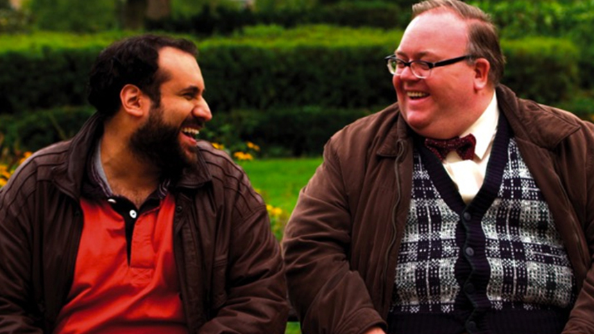 A screenshot of two mature heavyset men laughing together on a park bench, one of them brown and bearded in casual layers and the other white and bespectacled in a cardigan and bow tie.